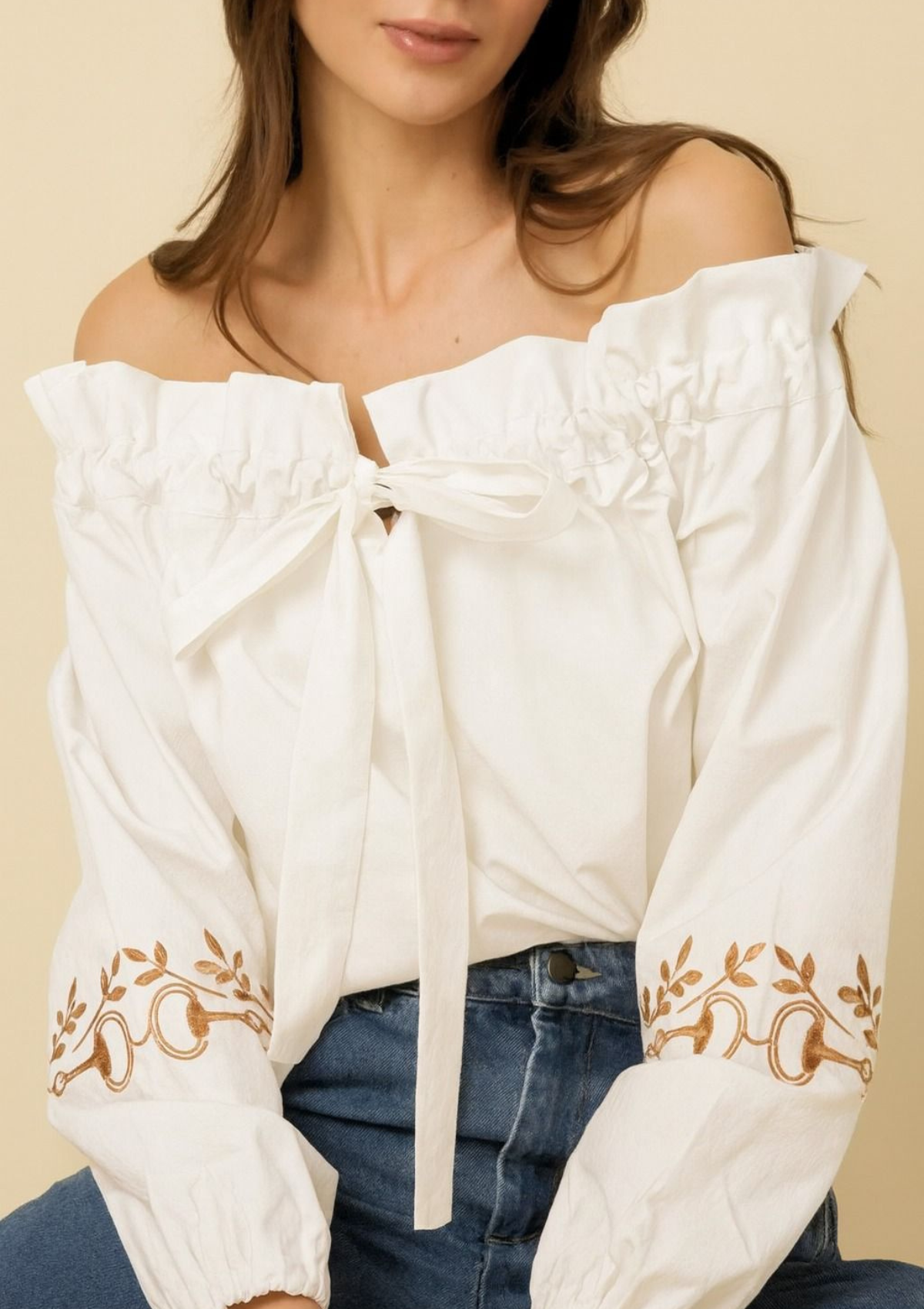 White off-shoulder blouse with floral embroidery on long sleeves, worn by a person against a beige background.