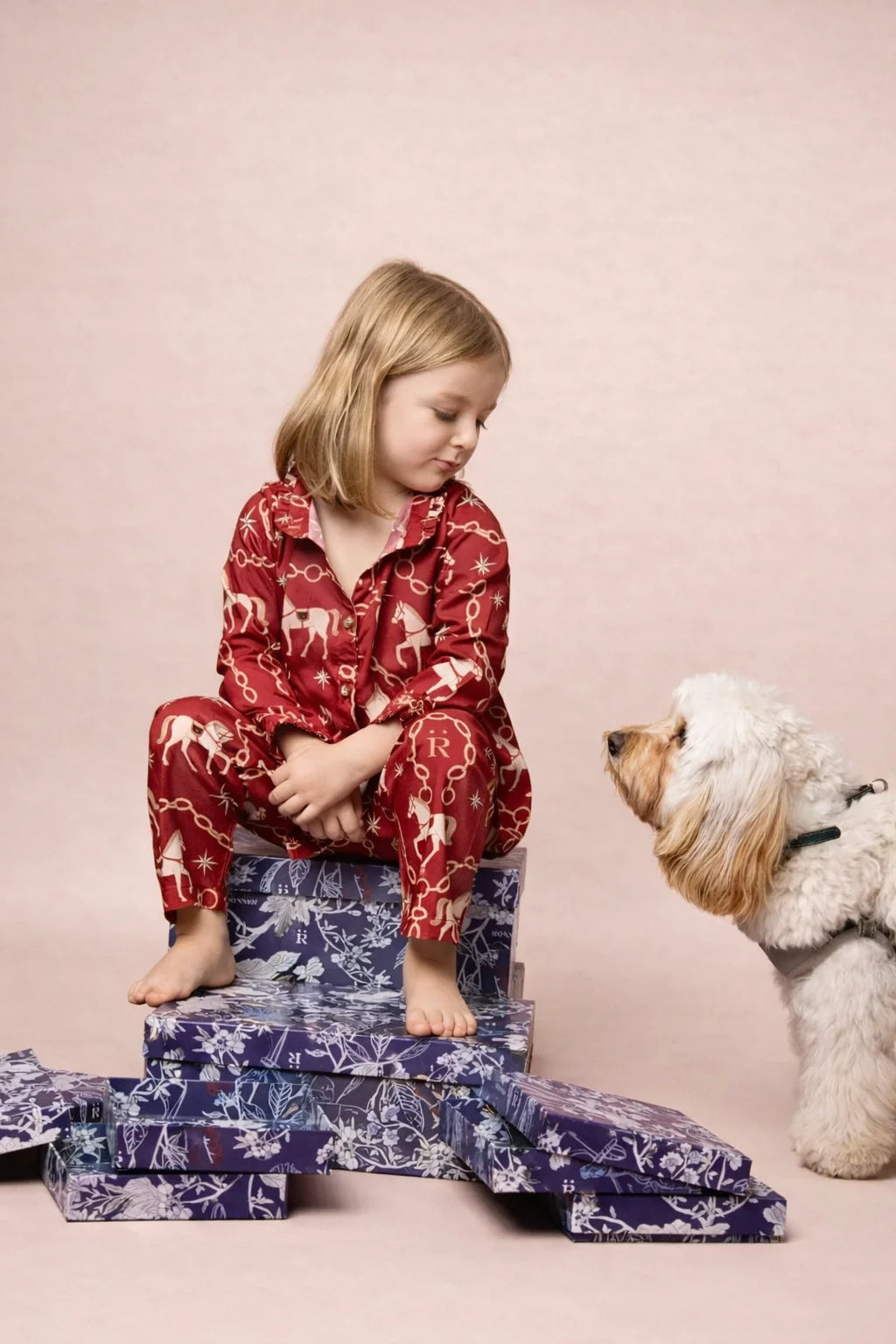 Child in red pajamas sitting on a stack of patterned boxes with a dog beside them on a pink background